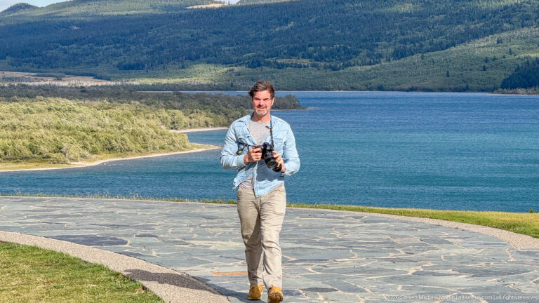 Visitor holding a camera on a stone overlook above Upper Waterton Lake, with forested hills beyond.