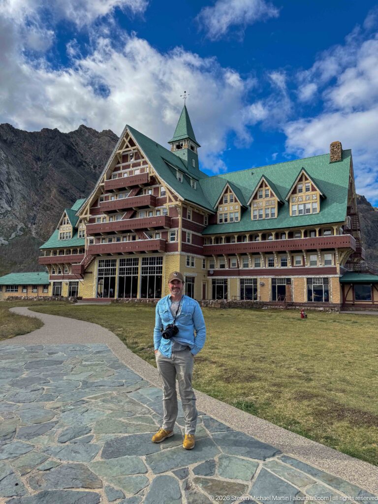 Visitor standing on a stone path before the Prince of Wales Hotel, with green roofs and rocky ridge behind.
