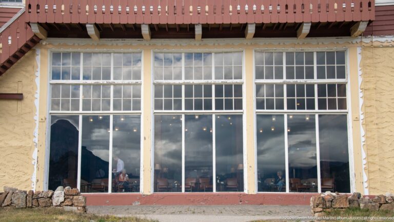 Tall multi-pane hotel windows reflecting clouds and mountains, set above a low rough-stone base beneath a red balcony.