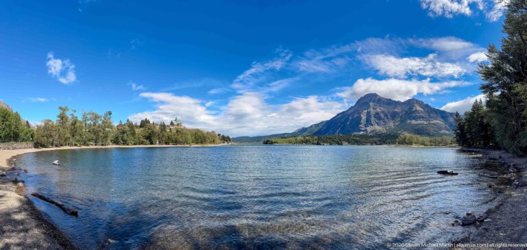 Upper Waterton Lake with a steep rocky mountain beyond wooded shorelines under blue sky and scattered clouds.