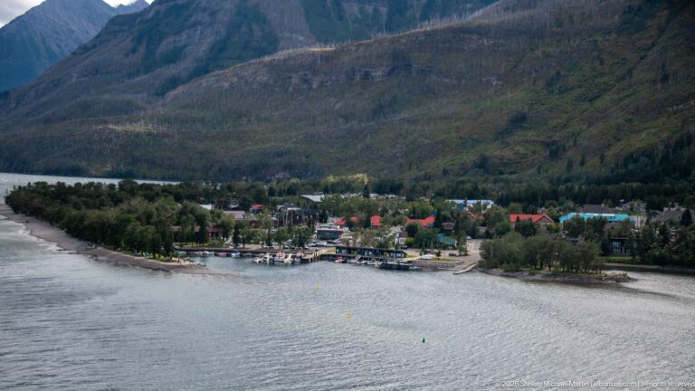Elevated view across water to Waterton townsite, docks, trees, and buildings beneath a broad mountain slope.