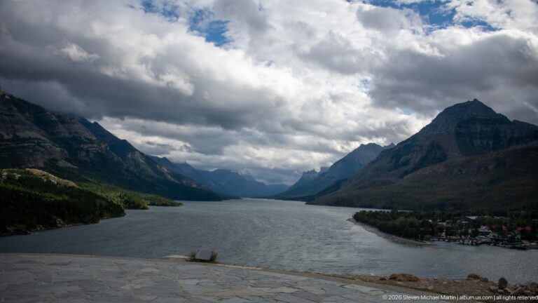 Upper Waterton Lake stretching between steep mountains beneath heavy clouds, viewed across a stone overlook with townsite at right.