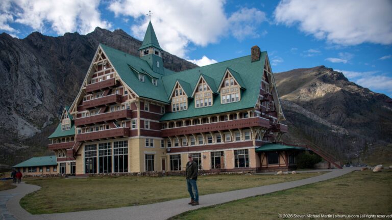 Visitor standing on a path beside the Prince of Wales Hotel, with tall gable, green roof, balconies, and mountain behind.