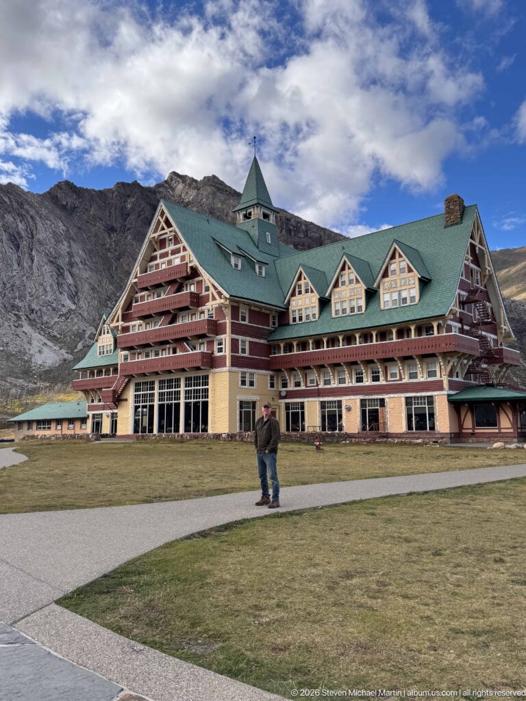 Wide view of Prince of Wales Hotel across a lawn, with a visitor on the path and rocky ridge behind.