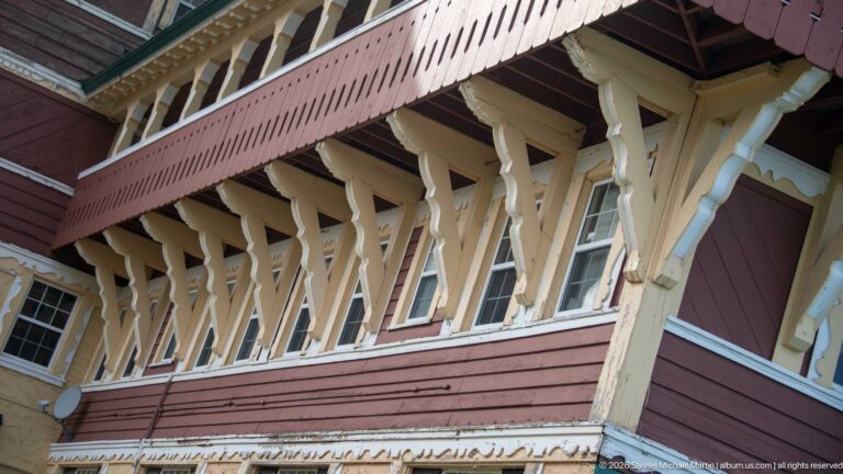 Close upward view of pale timber balcony brackets, red siding, windows, white pipes, and decorative trim on the hotel.