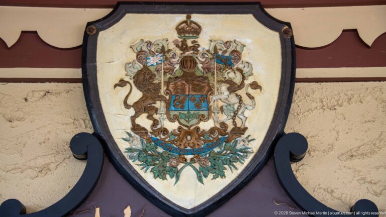 Weathered shield-shaped heraldic emblem with crown, supporters, and colorful arms mounted on the hotel exterior.