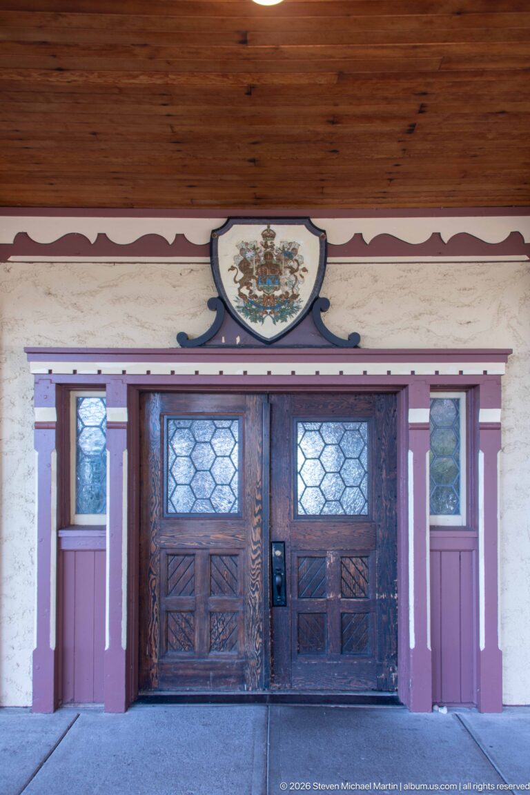 Hotel entrance with dark double doors and leaded glass beneath a shield-shaped heraldic emblem and wood-paneled soffit.