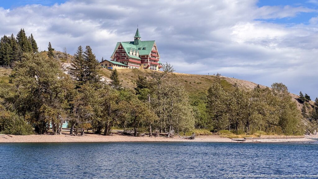 Prince of Wales Hotel rising above a wooded bluff across blue water, with its green roof visible against clouds.
