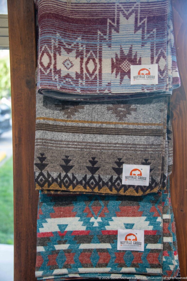 Three folded patterned textiles stacked on a wooden display, with red, turquoise, brown, and cream geometric designs.
