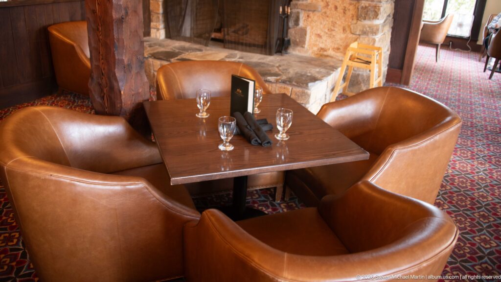 Four brown leather chairs around a wooden table beside a stone fireplace on red patterned carpet.