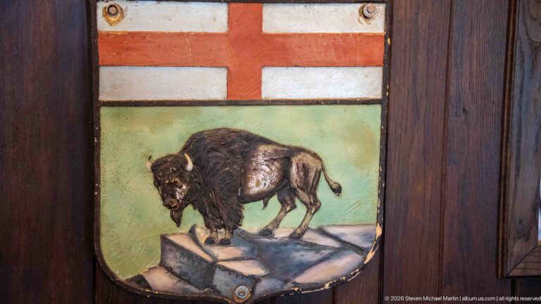 Painted shield showing a bison on rocks beneath a red cross, mounted on dark wooden paneling.