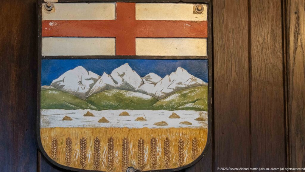 Painted shield with snowy mountains, green foothills, and a golden wheat field beneath a red cross.
