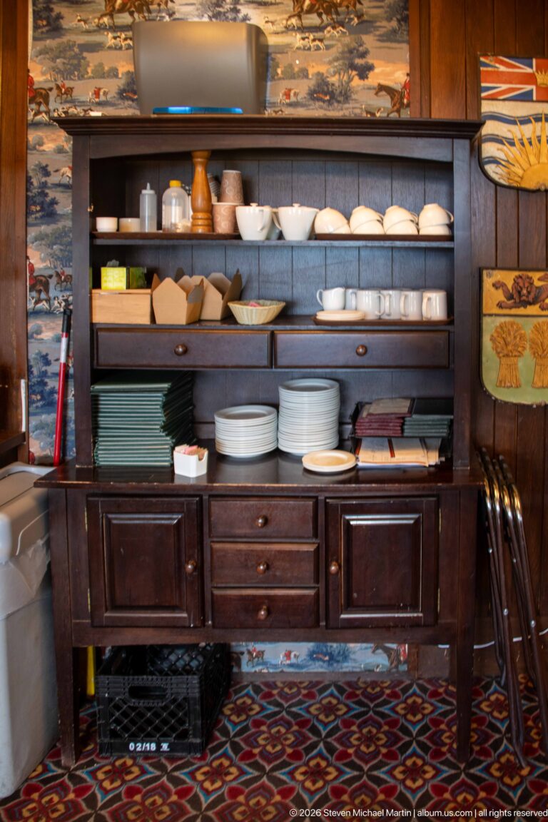 Dark wooden service cabinet filled with stacked white plates, bowls, cups, and trays above patterned carpet.