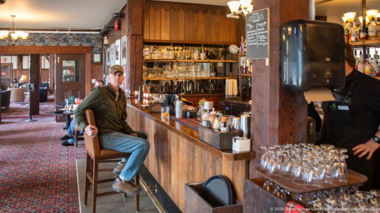 Patron seated at a wooden hotel bar while staff work behind the counter among bottles and glassware.