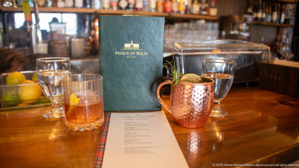 Green Prince of Wales menu beside a copper cocktail mug, water glasses, and printed menu on a wooden bar.