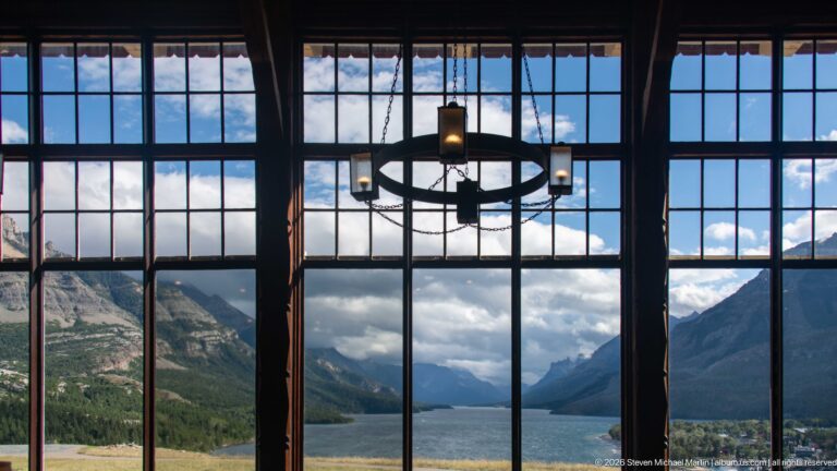 Tall lobby windows frame Upper Waterton Lake and mountains, with a circular chandelier silhouetted against the bright view.