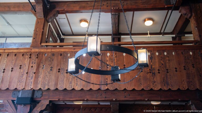 Circular iron chandelier with rectangular lanterns hanging in front of a carved wooden balcony.