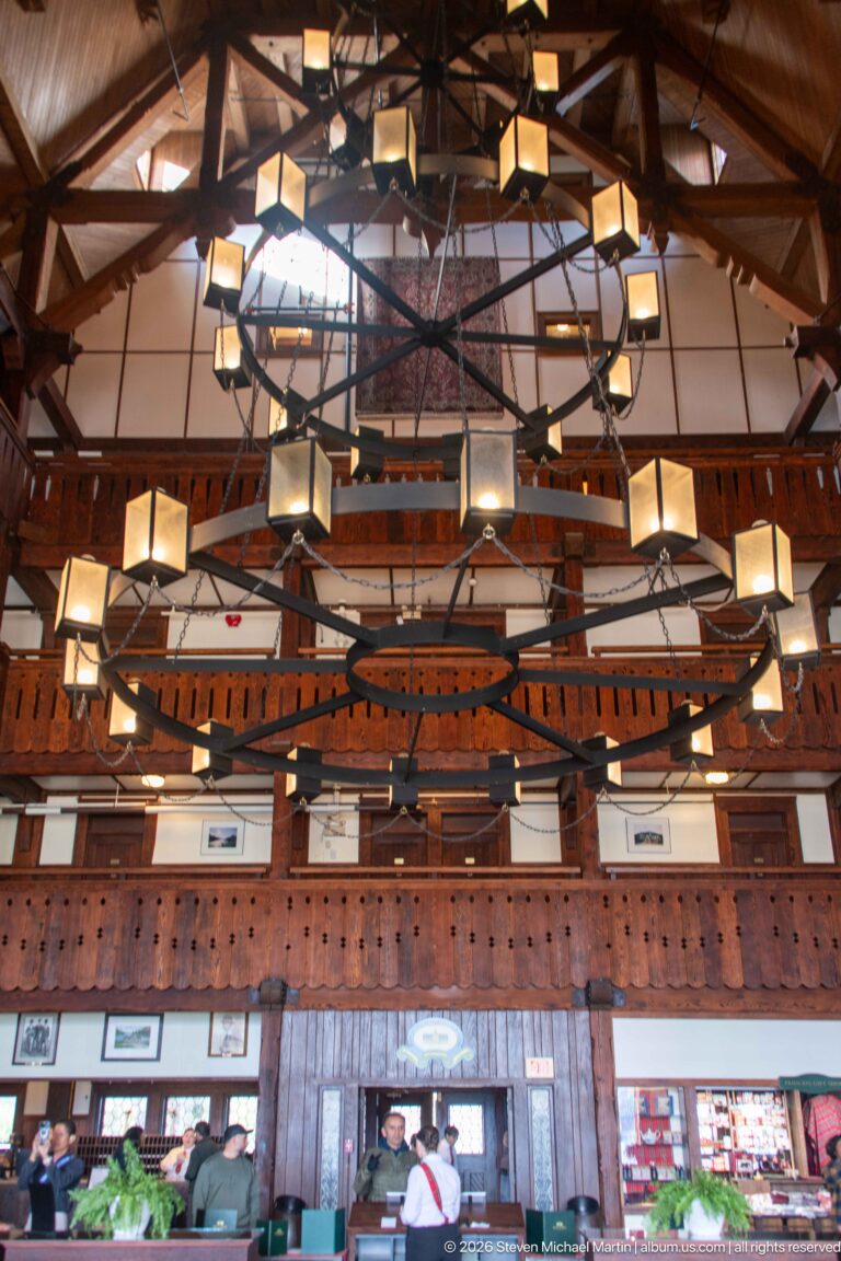 Large circular chandelier above the Prince of Wales Hotel lobby, with guests, seating, and carved wooden balconies below.