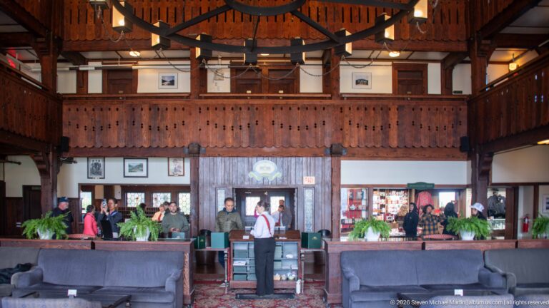 Guests stand in a timber-paneled hotel lobby beneath a carved balcony, with upholstered seating and ferns in the foreground.