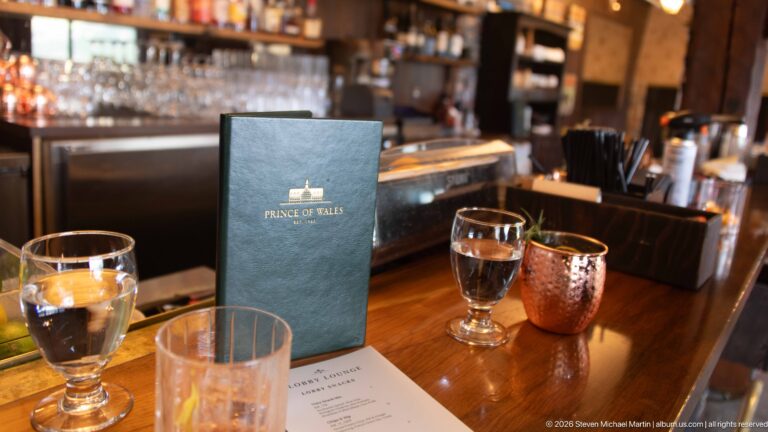 Prince of Wales menu, water glasses, copper mug, and lounge sheet arranged on a polished wooden bar.