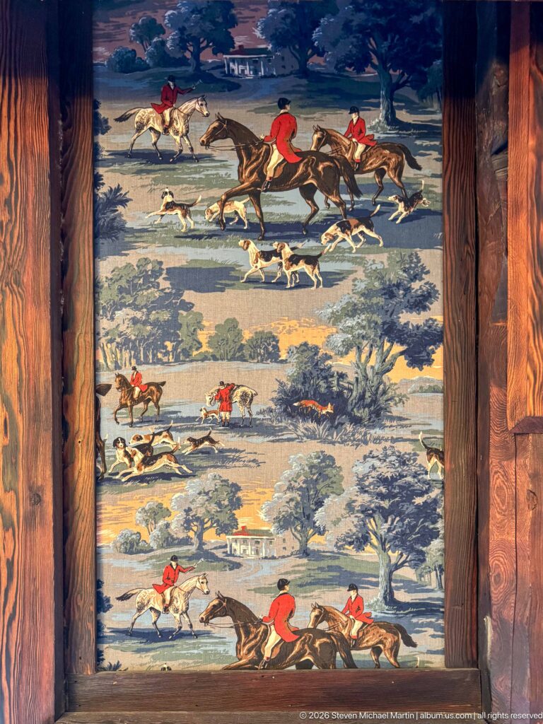 Framed blue-green textile patterned with red-coated riders on horseback and packs of hounds in hunting scenes.