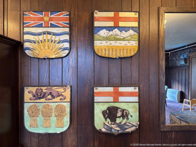 Four painted shields on dark wood showing a sunburst and waves, mountains and wheat, wheat sheaves, and a bison.