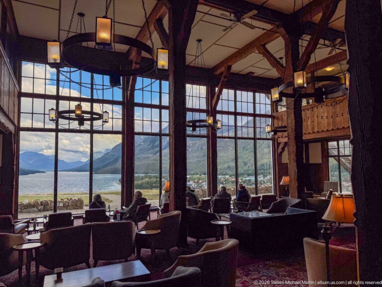 Hotel lobby seating faces tall windows overlooking Upper Waterton Lake and mountains, with guests silhouetted against the view.