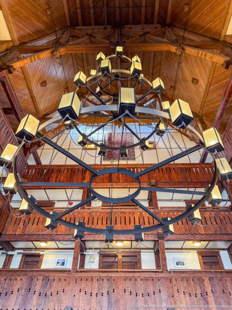 Low-angle close view of a huge circular iron chandelier with box lanterns beneath a warm wooden ceiling.