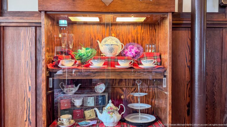 Glass cabinet displaying teapots, cups, saucers, tea packages, and serving stands on red tartan fabric.
