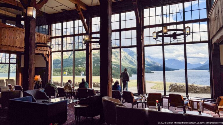 Guests in the timber lobby look through tall windows toward Upper Waterton Lake and surrounding mountains.