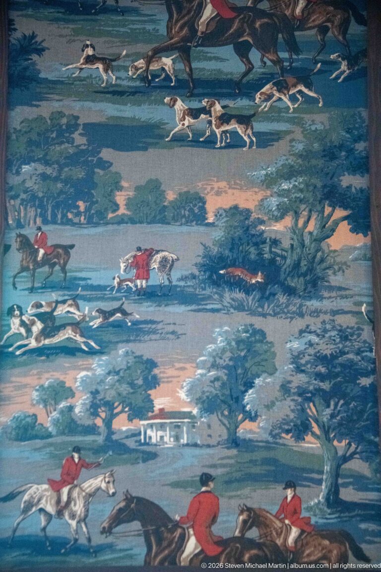 Close detail of blue-green textile with red-coated riders, galloping horses, white hounds, trees, and hunting scenes.