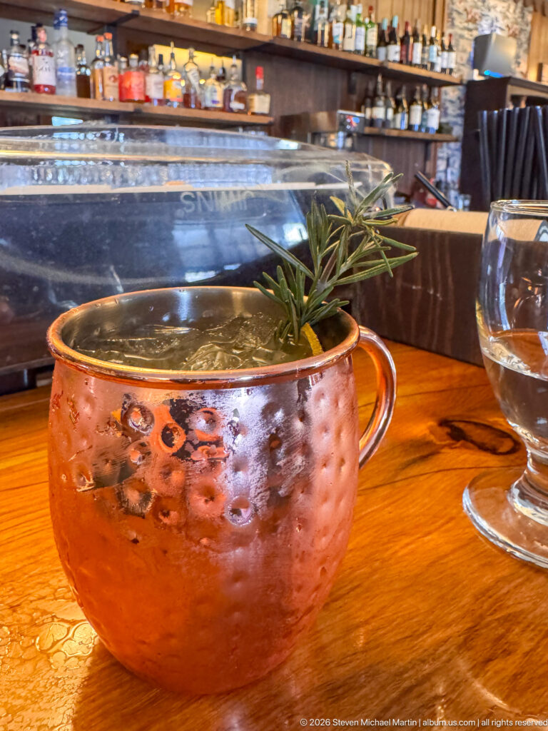 Hammered copper mug holding an iced drink with a rosemary sprig on a polished wooden bar.