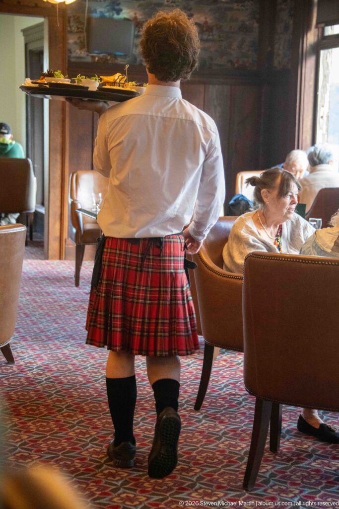 Server seen from behind wearing a white shirt, red tartan kilt, dark knee socks, and black shoes in the lounge.