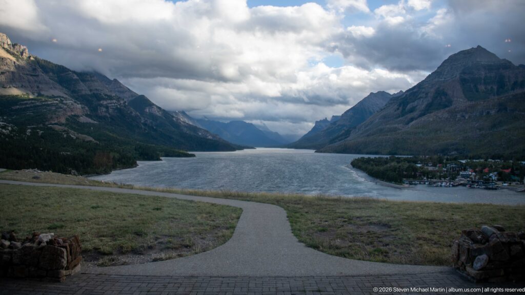 Path descending from a grassy bluff toward Upper Waterton Lake, mountains, clouds, and the Waterton townsite.