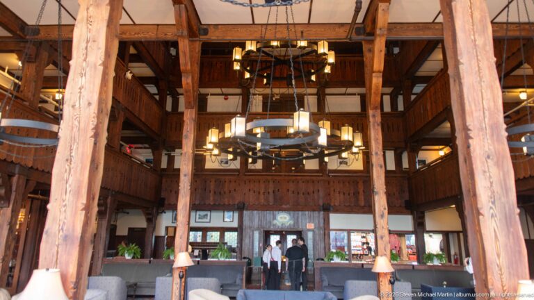 Tall timber columns frame a circular chandelier, balconies, reception area, and seating in the Prince of Wales Hotel lobby.