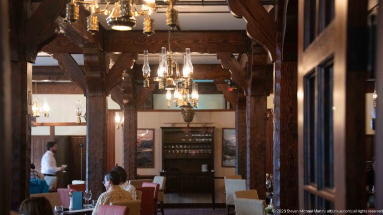 Timber-framed dining room with chandeliers, set tables, upholstered chairs, and a few seated guests.