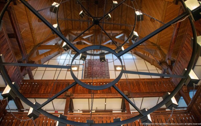 Large black circular chandelier with radial metal supports and square lanterns beneath exposed timber roof framing.