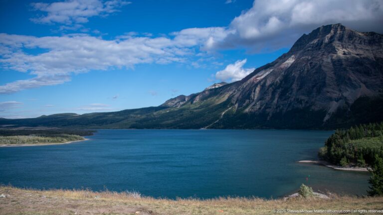 Elevated view of Upper Waterton Lake curving below a massive rocky mountain with forested lower slopes.
