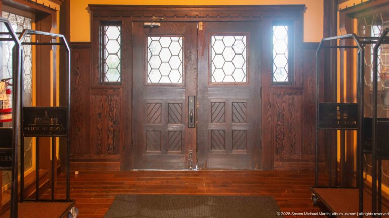 Wooden double doors with geometric leaded-glass panes, sidelights, diagonal lower panels, and dark timber trim.