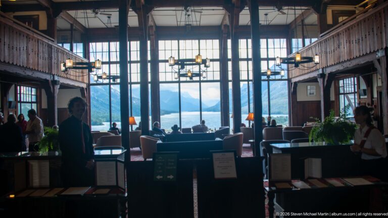 Hotel lobby with guests and seating silhouetted against tall windows overlooking Upper Waterton Lake and mountains.