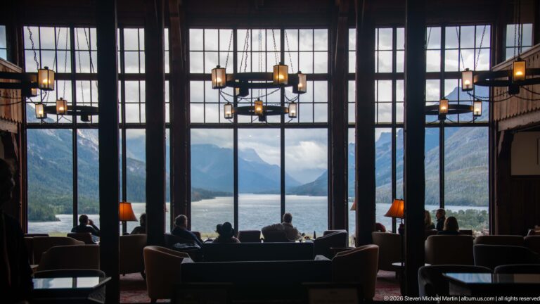 Seated guests face tall lobby windows framing Upper Waterton Lake between steep mountain slopes.