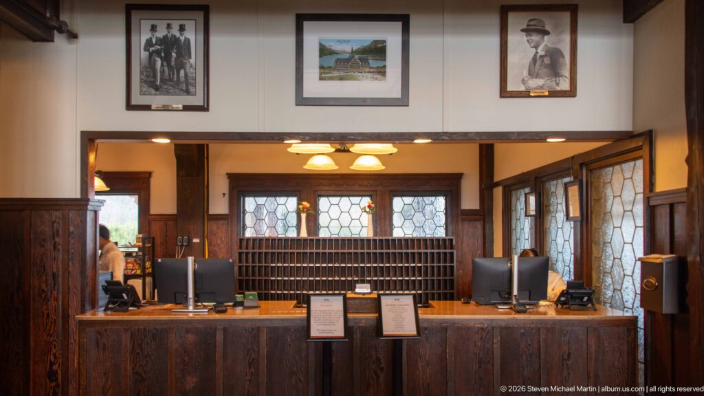 Wood-paneled hotel reception desk with computers, framed pictures, leaded glass, and a wall of small numbered slots.