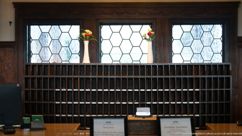 Rows of narrow wooden hotel cubbies beneath geometric leaded-glass windows and two small flower vases.