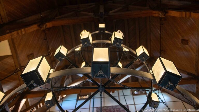 Upward view of square lanterns arranged around a black circular chandelier beneath a wood ceiling.