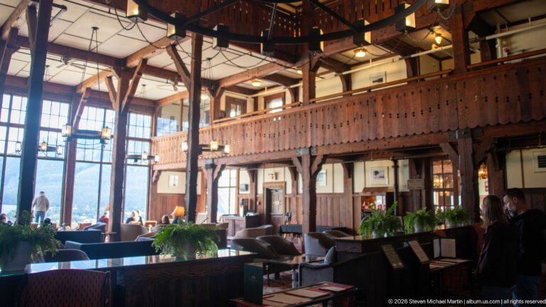 Wide lobby view with sofas, plants, hanging lights, timber balconies, and tall windows at left.