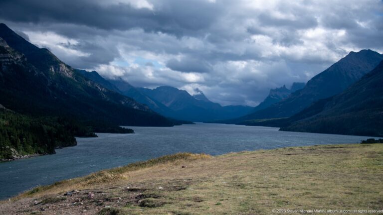 Upper Waterton Lake extending between dark mountain slopes beneath dramatic clouds, viewed from a grassy bluff.