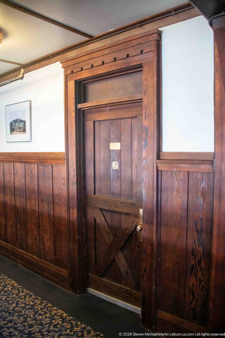 Hotel corridor with dark wood wainscoting, patterned carpet, framed art, and a solid paneled guest-room door.
