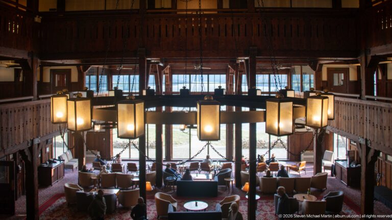 Elevated view over lobby tables and seating beneath a large chandelier, framed by timber balconies and tall windows.