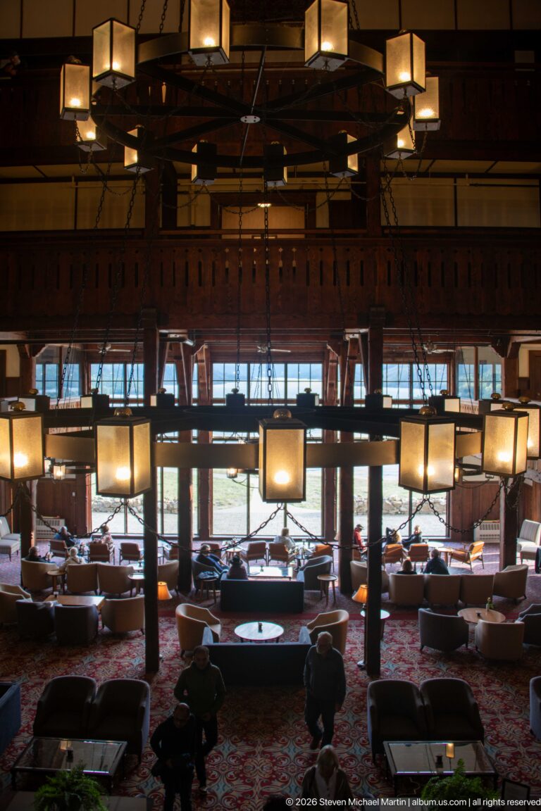 Upper-level view of lounge seating, patterned carpet, and a broad chandelier beneath a dark timber balcony rail.