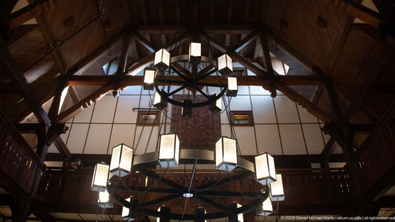 Exposed timber roof trusses rise above a large circular chandelier with square lanterns in the hotel lobby.